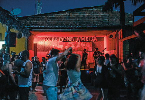 Imagem: Fotografia. Uma banda sobre um palco e na frente deles, várias pessoas estão dançando. Fim da imagem.