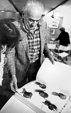 Imagem: Fotografia em preto e branco. Carybé, homem com cabelo penteado para trás, de camisa e terno, está olhando um livro grande.  Fim da imagem.