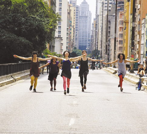 Imagem: Fotografia. Cinco pessoas estão andando em uma ponte com os braços abertos. Ao fundo, pessoas, árvores e prédios. Fim da imagem.