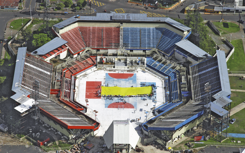 Imagem: Fotografia. Vista aérea de uma arena grande com formato de octógono. Em volta há arquibancadas vermelhas e azuis. Ao redor há árvores e construções. Fim da imagem.