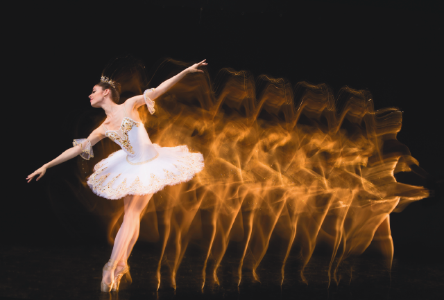 Imagem: Fotografia. Uma mulher com cabelo preso, tiara, vestido e sapatilhas de bailarina está nas pontas dos pés e com os braços abertos. Atrás dela, sombra de seus movimentos. Fim da imagem.