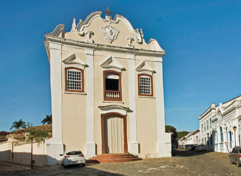 Imagem: Fotografia. Construção com dois andares, paredes bege com detalhes brancos. Janelas e porta marrons. Em volta há casas e carros. Fim da imagem.