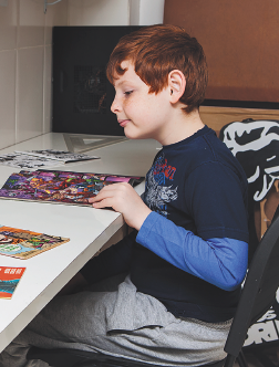 Imagem: Fotografia. Um menino ruivo com camiseta azul e calça cinza está sentado e lendo uma revista em quadrinhos, que está em cima de uma mesa. Fim da imagem.