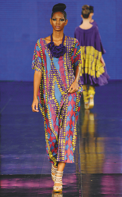 Imagem: Fotografia. Uma modelo com cabelo preso, colar grande e roxo, vestido colorido e sandálias marrons está andando em uma passarela. Ao fundo, uma modelo com vestido roxo e amarelo está de costas e se afastando. Fim da imagem.