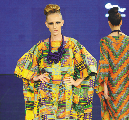 Imagem: Fotografia. Uma modelo loira com cabelo preso, colar grande e roxo e vestido colorido com mangas compridas está com as mãos na cintura. Ao fundo, uma modelo com vestido verde e laranja está de costas e se afastando. Fim da imagem.