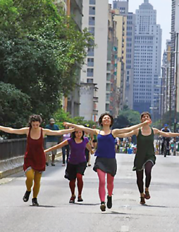 Imagem: Fotografia. Quatro mulheres estão andando com os braços abertos. Ao fundo, prédios e árvores. Fim da imagem.