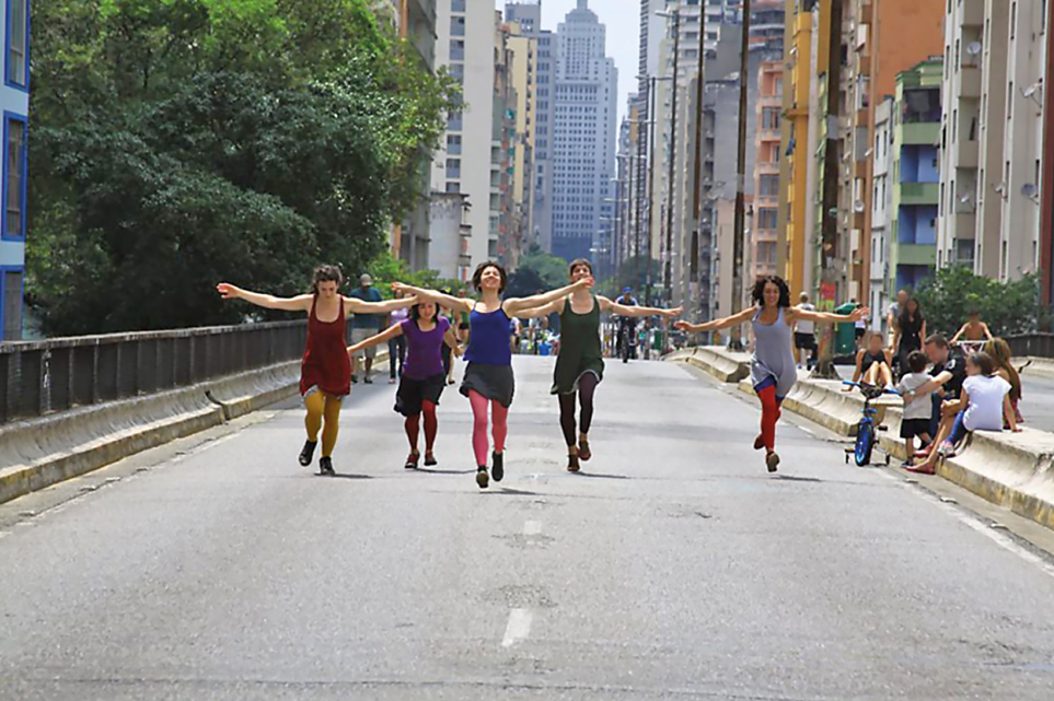 Imagem: Fotografia. Quatro mulheres estão andando em um viaduto com os braços abertos. Ao fundo, prédios e árvores. Fim da imagem.