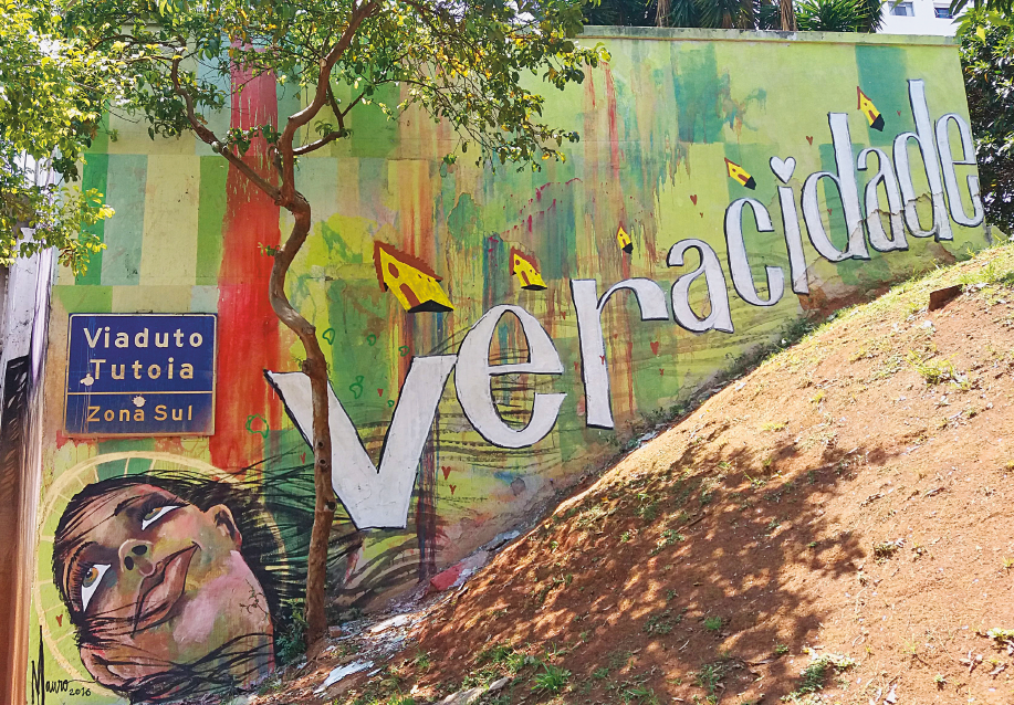 Imagem: Fotografia. Um muro pintado de verde. No centro, a palavra “VERACIDADE” e acima da palavra há algumas casas amarelas. À esquerda, rosto de uma jovem olhando para cima, onde há uma placa com a informação: VIADUTO TUTOIA – ZONA SUL. Ao lado há uma avenida e na frente, árvores. Fim da imagem.