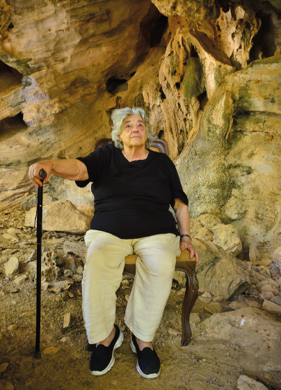 Imagem: Fotografia. Niède, senhora com cabelo grisalho, camiseta e sapatos pretos e calça bege está sentada em uma cadeira, dentro de uma caverna, olhando para cima e com a mão direita apoiada em uma bengala.  Fim da imagem.