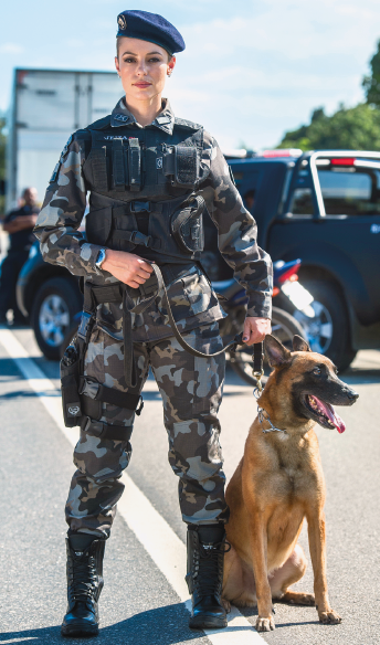 Imagem: Fotografia. Mulher com uniforme militar e quepe está segurando a coleira de um cachorro marrom, que está sentado ao seu lado. Ao fundo, carros e árvores. Fim da imagem.