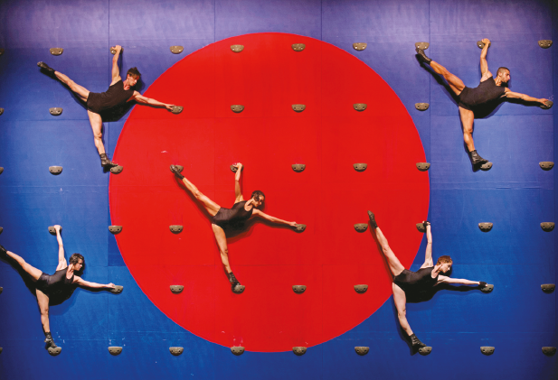 Imagem: Fotografia. Cinco bailarinos com roupa preta estão com as pernas direitas esticadas para cima e os braços abertos. Ao fundo há várias bolinhas sobre um círculo vermelho em um retângulo azul. Fim da imagem.