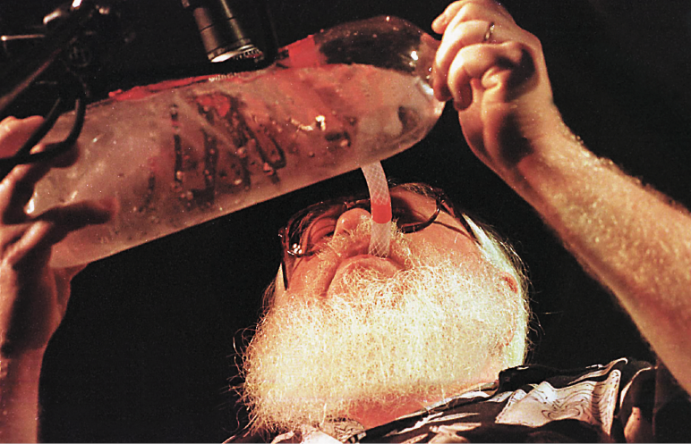 Imagem: Fotografia. Vista de baixo de Hermeto com um cano na boca, que está conectado a uma garrafa com água. Fim da imagem.