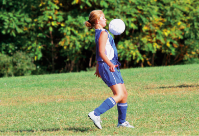 Imagem: Fotografia. Menina de cabelo curto loiro, vestindo colete e bermuda azul. Está com uma bola em movimento na direção do tronco. Fim da imagem.