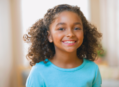 Imagem: Fotografia. Menina de cabelo cacheado castanho, sorrindo. Veste camisa azul. Fim da imagem.