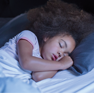 Imagem: Fotografia. Menina de cabelo longo crespo, vestindo camiseta branca. Está deitada em uma cama, dormindo.  Fim da imagem.