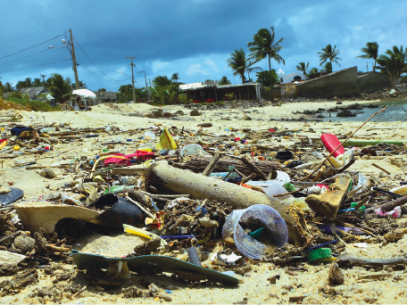 Imagem: Fotografia. Areia da praia com lixo e resíduos diversos por toda a praia. Fim da imagem.