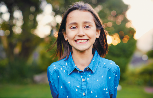 Imagem: Fotografia. Menina de cabelo curto liso castanho, sorrindo. Veste camisa azul com bolinhas brancas. Fim da imagem.