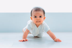 Imagem: Fotografia. Menino bebê de cabelo curto preto, vestindo roupinha branca. Fim da imagem.
