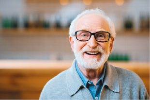 Imagem: Fotografia. Homem idoso de cabelo curto e barba curta grisalha, e óculos de armação quadrada preta, vestindo camisa azul e casaco verde. Fim da imagem.