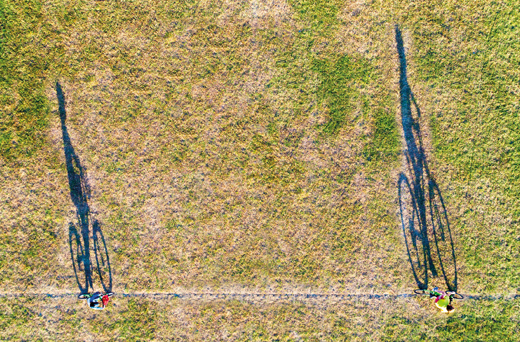 Imagem: Fotografia complementar das páginas 64 e 65. Vista aérea de campo gramado com fileira de terra onde há dois ciclistas andando com sombra ampliada à esquerda. Fim da imagem.