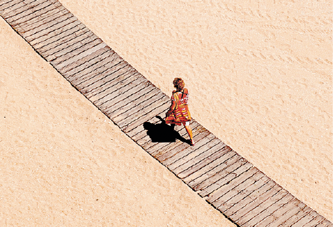 Imagem: Fotografia. Vista aérea de mulher andando em uma passarela de madeira sobre areia. Sua sombra está diminuída abaixo dela. Fim da imagem.