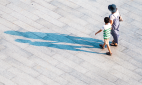Imagem: Fotografia. Vista aérea de mulher e menino de mãos dadas andando em uma calçada. À esquerda, sombra ampliada dos dois.  Fim da imagem.