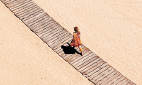 Imagem: Fotografia. Vista aérea de mulher andando em uma passarela de madeira sobre areia. Sua sombra está diminuída abaixo dela. Fim da imagem.