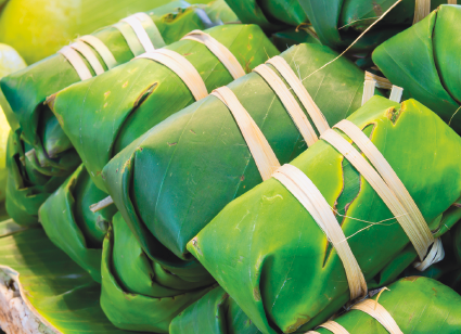 Imagem: Fotografia. Embalagens fechadas empilhadas de folhas de bananeira com fios de cipó. Fim da imagem.