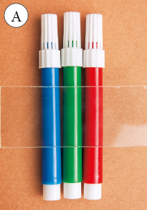 Imagem: Fotografia. Três canetas nas cores azul, verde e vermelho respectivamente. A: um pedaço de vidro retangular sobre a imagem.  Fim da imagem.
