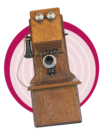 Imagem: Fotografia de um telefone comprido de madeira indicando telefone do início de 1900. Fim da imagem.