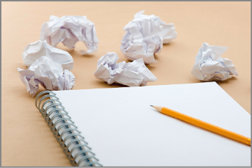 Imagem: Fotografia. Caderno com um lápis amarelo. Ao lado, há bolinhas de papel amassadas. Fim da imagem.