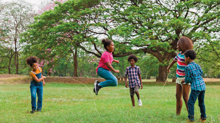 Imagem: Fotografia. Crianças brincam em um gramado de pular corta. Há duas meninas segurando a corda enquanto uma menina pula. Há dois meninos ao redor. Fim da imagem.