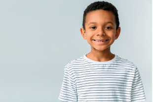 Imagem: Fotografia. Menino de cabelo curto preto, vestindo camiseta branca com listras pretas, sorrindo. Fim da imagem.