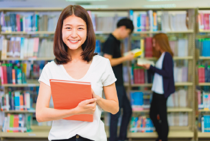 Imagem: Fotografia. Menina adolescente de cabelo longo castanho, vestindo camiseta branca. Está segurando um livro, está no interior de uma biblioteca. Fim da imagem.