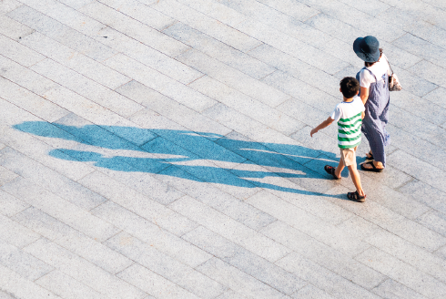 Imagem: Fotografia. Vista aérea de mulher e menino de mãos dadas andando em uma calçada. À esquerda, sombra ampliada dos dois.  Fim da imagem.