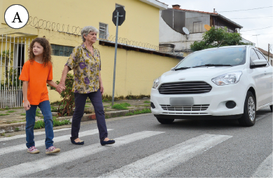 Imagem: Fotografia. A: Menina de mãos dadas com uma mulher idosa, atravessando a sua em uma faixa de pedestres. Ao lado, um carro parado aguarda.  Fim da imagem.