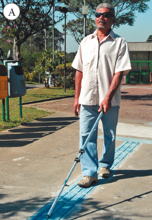 Imagem: Fotografia. A: homem de cabelo curto grisalho e óculos de sol preto, vestindo camisa bege e calça azul. Está segurando uma bengala, apoiada sobre faixa azul com elevações retas para guia.  Fim da imagem.