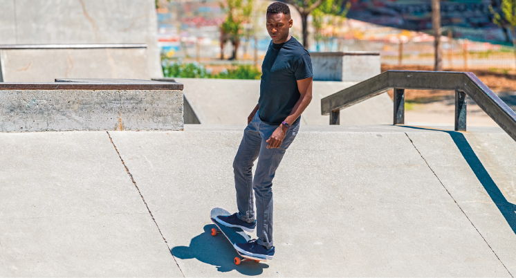 Imagem: Fotografia. Homem de cabelo curto preto, vestindo camiseta azul e calça cinza. Está andando de skate em uma pista com obstáculo durante o dia com sombra diminuída abaixo do homem. Fim da imagem.