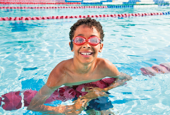 Imagem: Fotografia. Menino de cabelo curto castanho e óculos de natação vermelho. Está em uma piscina, apoiado sobre a divisória de setores da piscina. Fim da imagem.