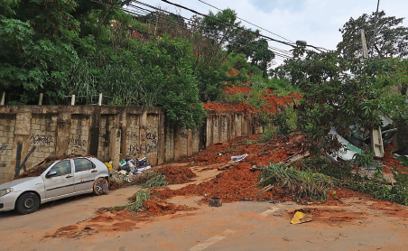 Imagem: Fotografia. Rua bloqueada por deslizamento de terra ultrapassando casas, árvores e objetos. Fim da imagem.