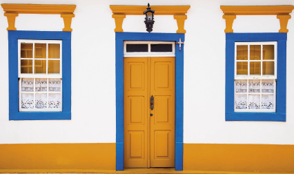 Imagem: Fotografia. Faixada de casa branca com faixas amarelas e contornos azuis nas portas e janelas. A porta é amarela. Fim da imagem.