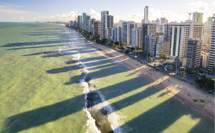 Imagem: Fotografia. Vista aérea de litoral com prédios formando suas sombras no mar de forma ampliada. Fim da imagem.
