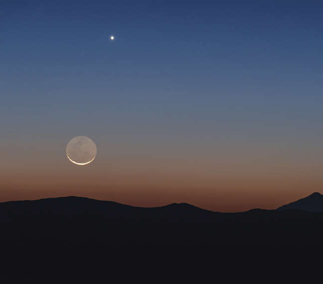 Imagem: Fotografia. Vista do horizonte iluminado pelo pôr-do-sol, deixando a terra sombreada. No cel há uma lua crescente e uma estrela. Fim da imagem.