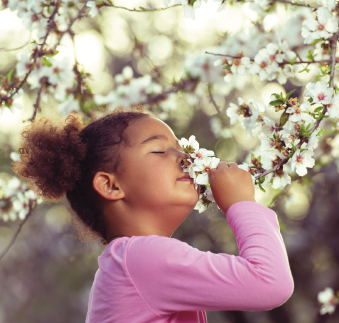 Imagem: Fotografia. Menina de cabelo curto cacheado castanho, vestindo camiseta rosa. Está cheirando uma flor. Fim da imagem.