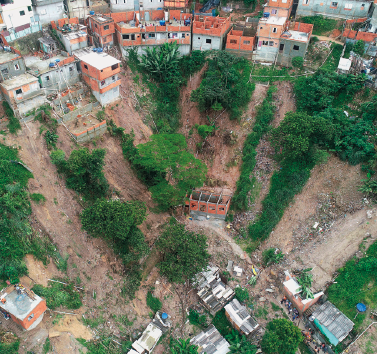 Imagem: Fotografia. Vista aérea de terra de um morro com deslizamento sobre casas irregulares. Fim da imagem.