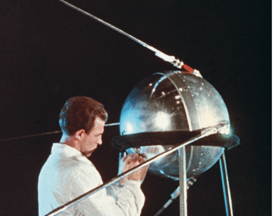 Imagem: Fotografia. Homem de cabelo curto castanho, vestindo casaco branco. Está ao lado de um satélite pequeno arredondado em uma estrutura metálica de suporte. Fim da imagem.