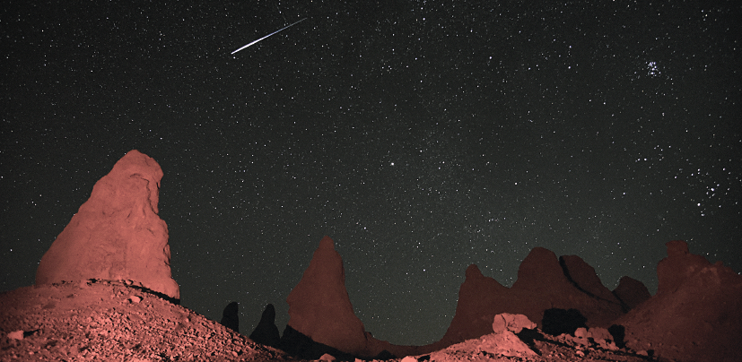 Imagem: Fotografia. Vista de deserto com rochas e área aberta. Sobre o céu escuro há estrelas e constelações. Fim da imagem.