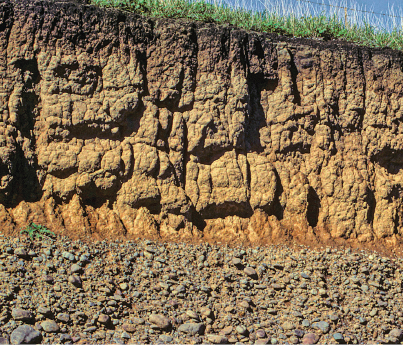 Imagem: Fotografia. Vista de perfil com rochas ligadas a terra com diferentes níveis de pedras. Na parte inferior há muitas pedras unidas. Na parte superior barro empedrado com rochas. Fim da imagem.