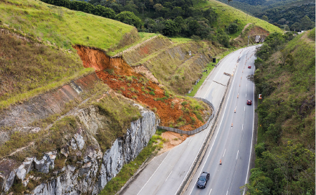 Imagem: Fotografia. Vista de montanha com deslizamento de rochas e terra sobre rodovia de seis vias, travando três vias da estrada. Fim da imagem.