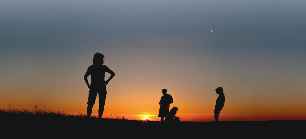 Imagem: Fotografia. Vista de pôr-do-sol com silhueta da sombra de quatro pessoas sobre o campo aberto sem árvores. Fim da imagem.